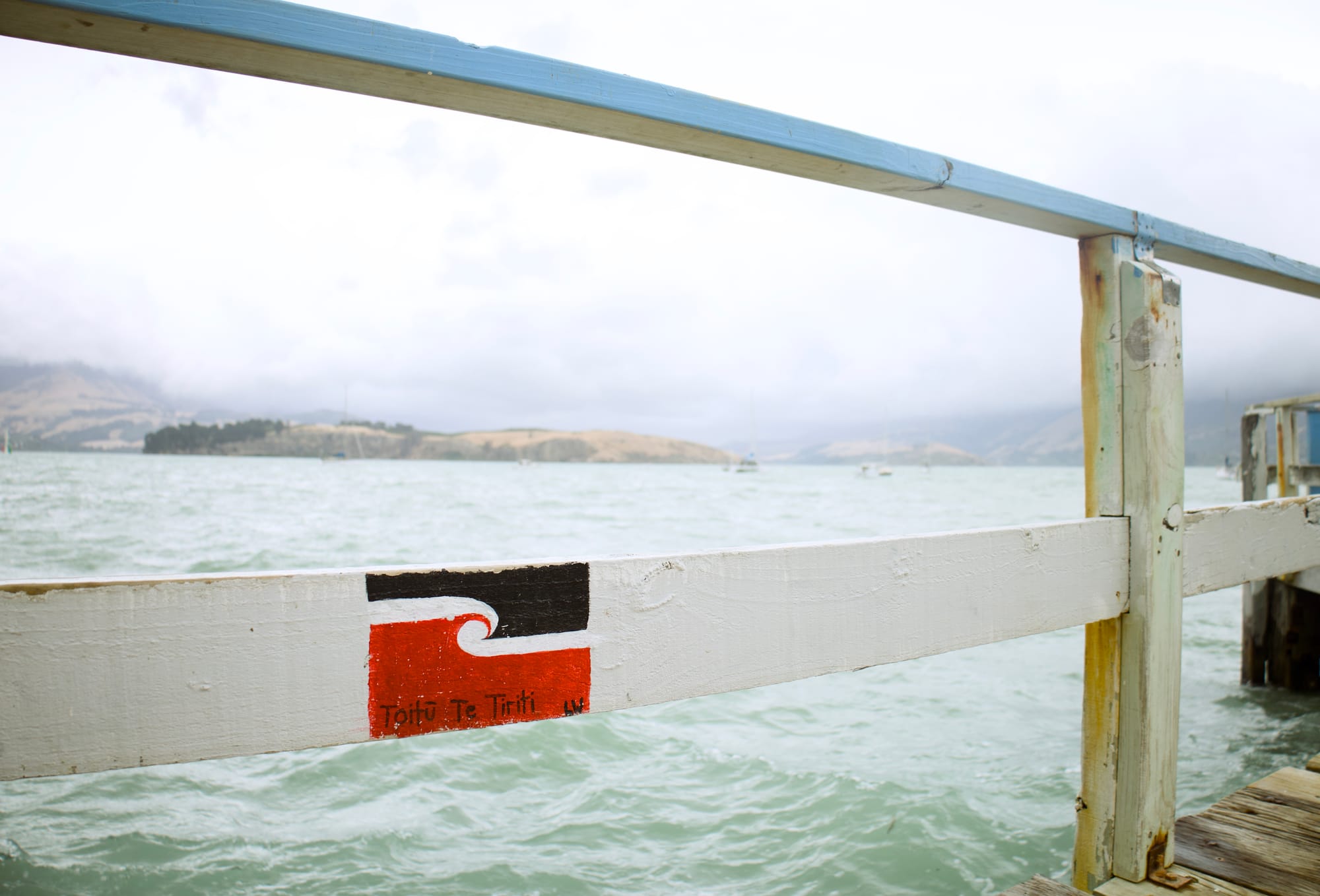A close up of railing on a dock, with the tino rangatiratanga flag painted on horizontal post. The sea in the background is choppy and the clouds are grey.