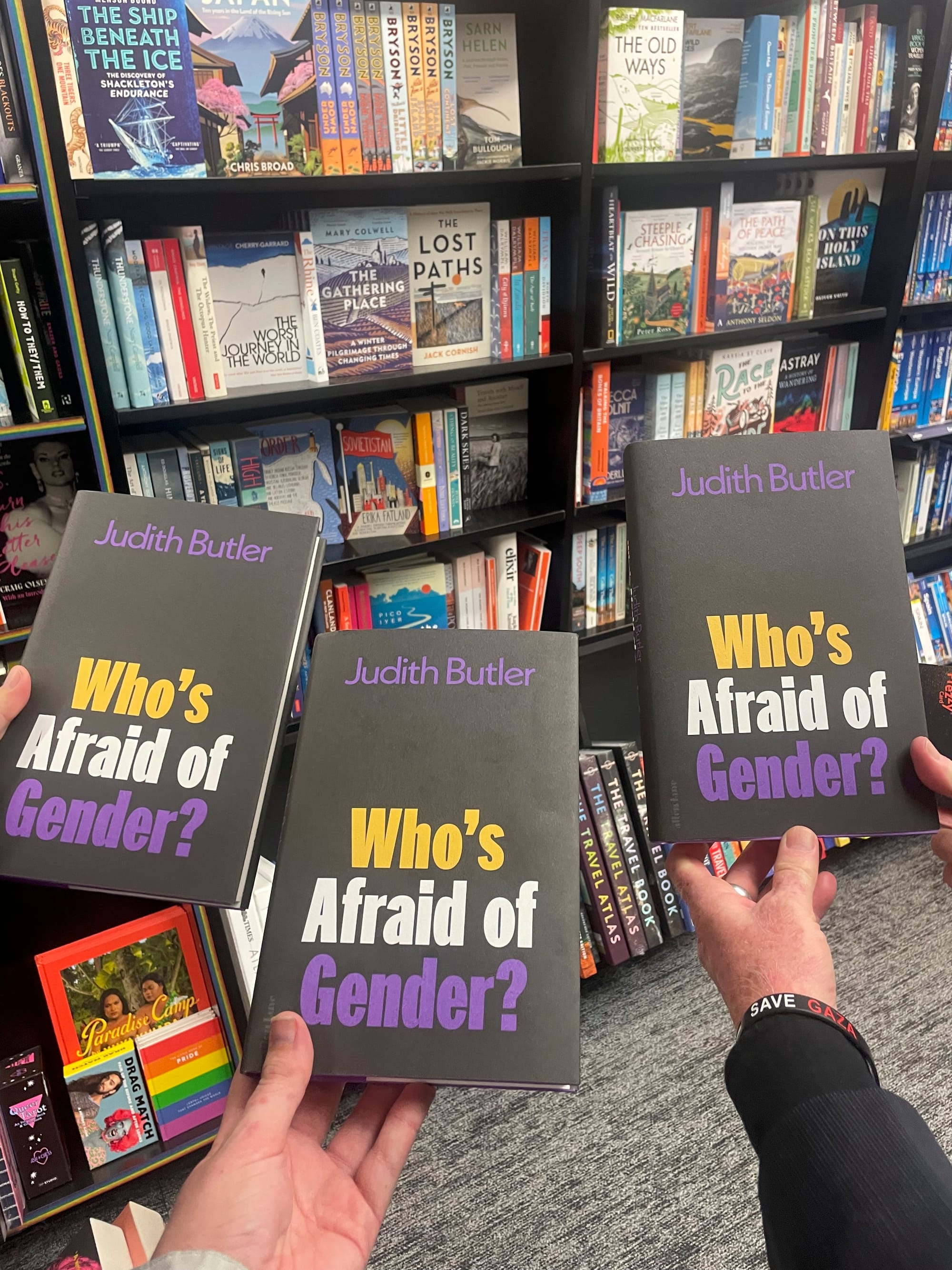 Three hands holding three copies of Who's Afraid of Gender against a backdrop of bookshelves.