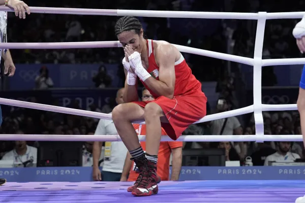 Imane Khelif after winning her quarterfinal, resting on the ropes around the ring, hands together, held to her face, smiling.